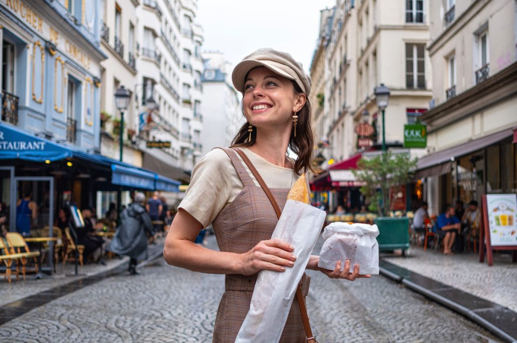 A woman with a baguette on her hand and a bag in another hand walking in the streets of Paris.
